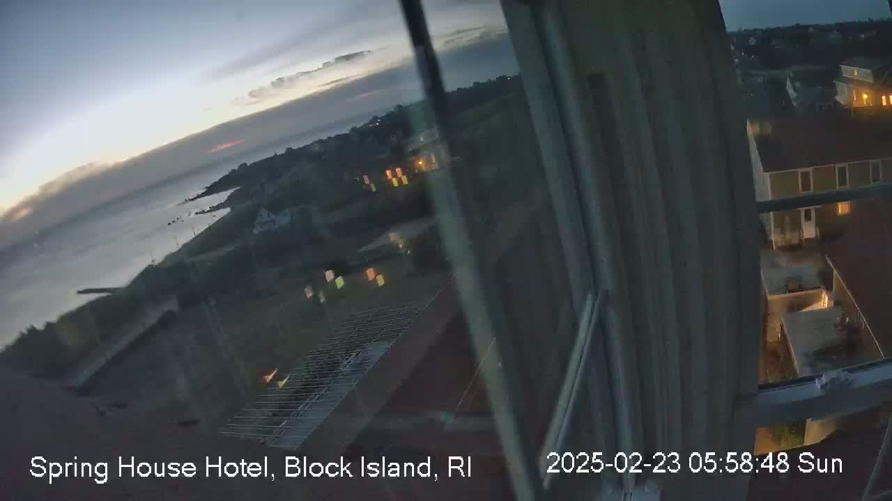 A twilight view from a window shows a calm ocean, a coastline with buildings, and a few illuminated windows in the surrounding neighborhood.