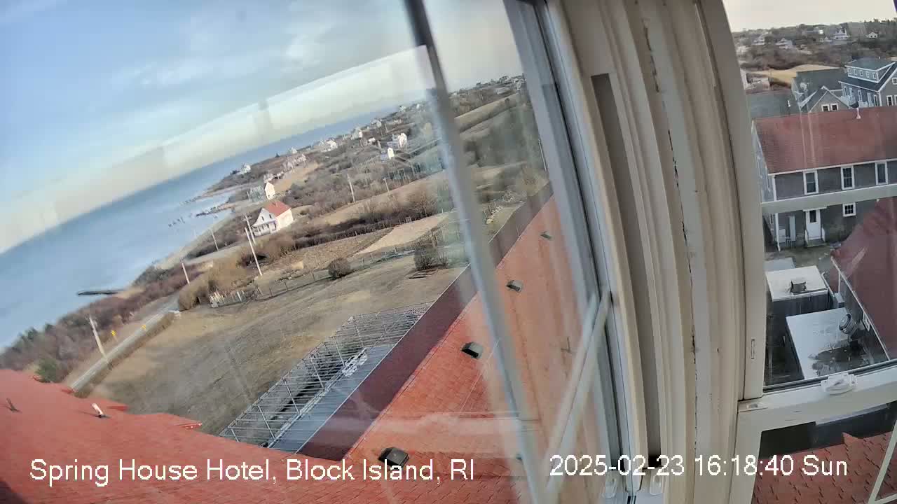 A sunny view of a coastal town and ocean from a window, showing red rooftops, bare winter trees, and a calm sea.