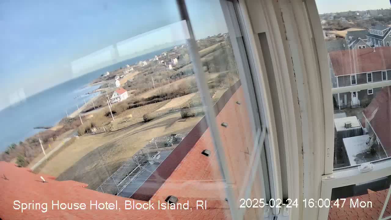 A view from a window shows a clear, sunny day overlooking a coastal town with a body of water, buildings, and open land.