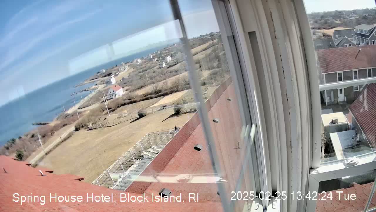 A view from a window shows a sunny coastal scene with houses, a body of water, and bare winter or early spring vegetation.