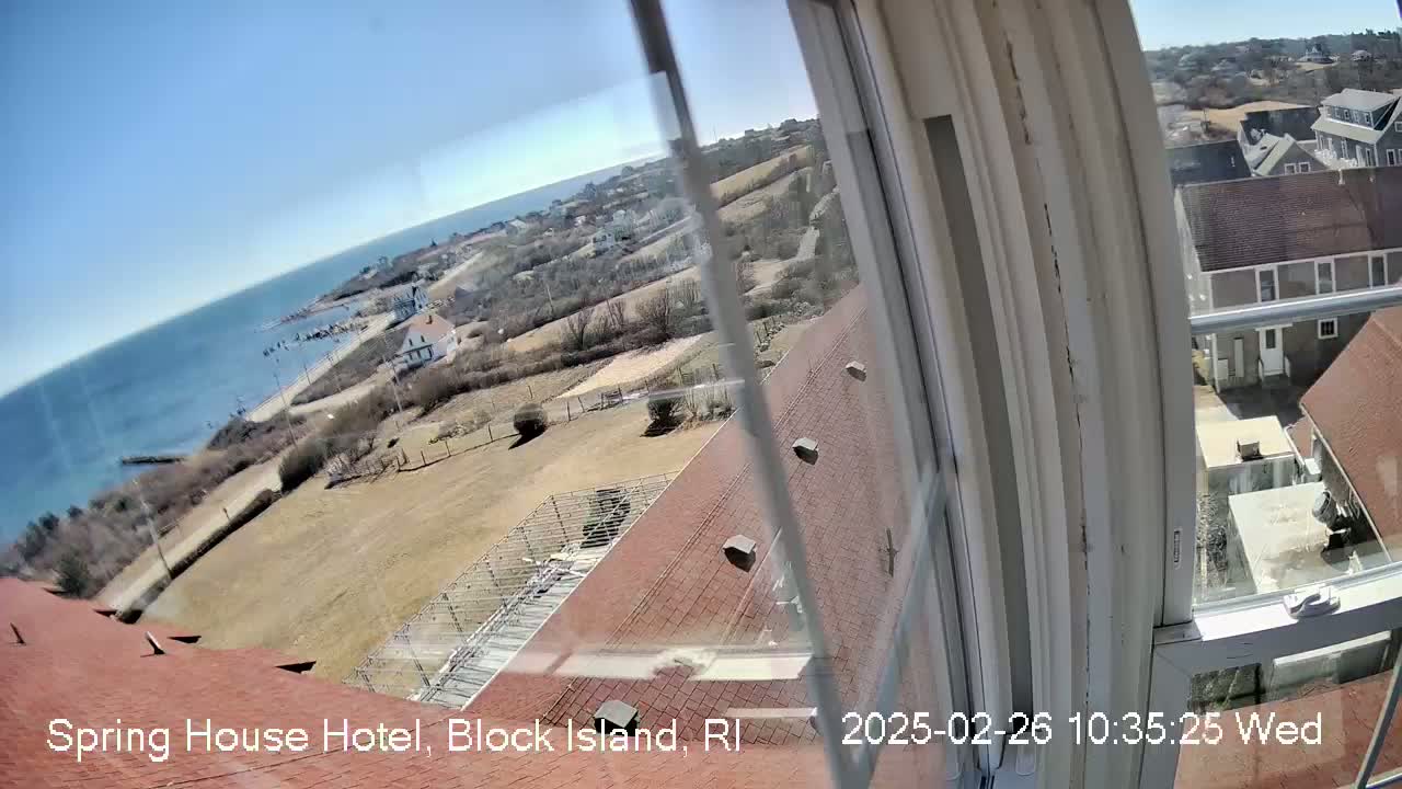 A view from a window shows a clear, sunny day overlooking a coastal town with a body of water and brown, sparsely vegetated land.