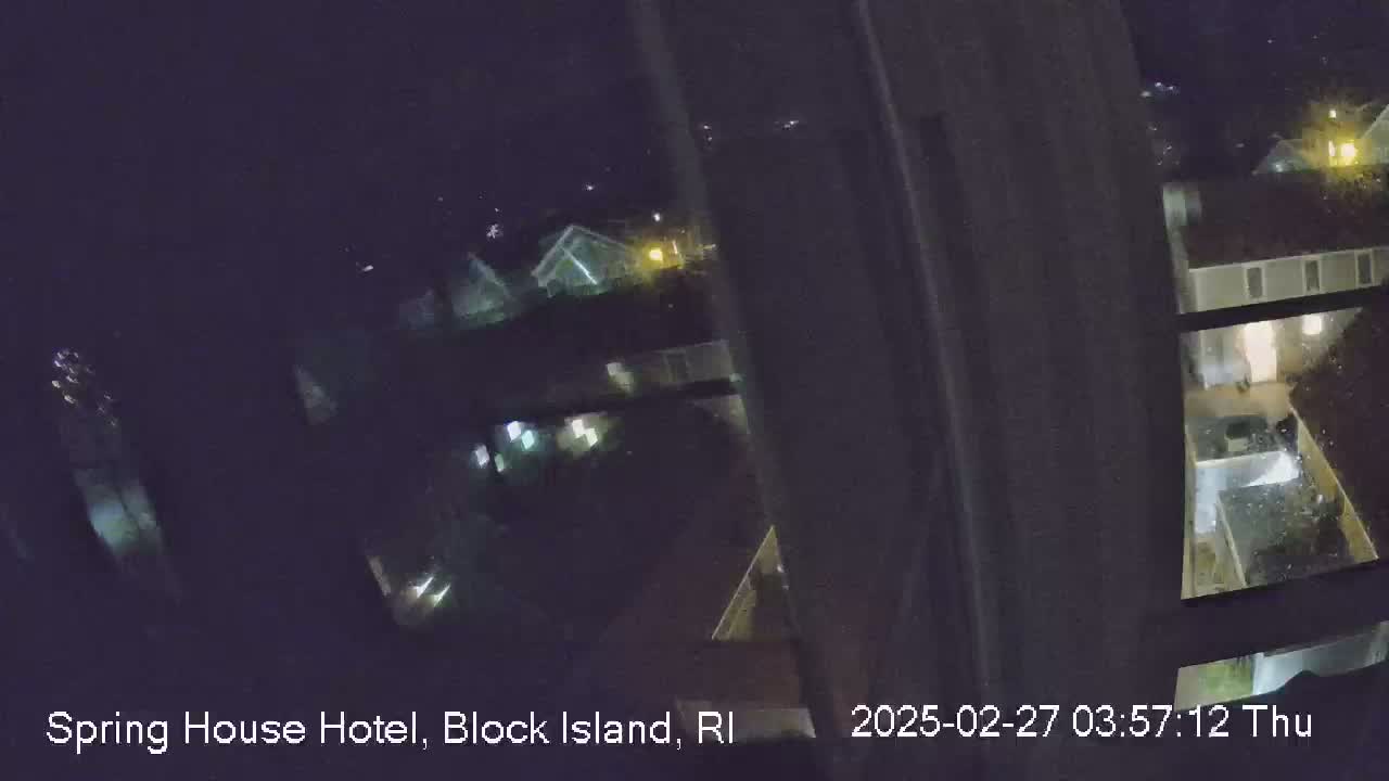 A nighttime view from a window shows several illuminated buildings and yards with rain or snow falling.