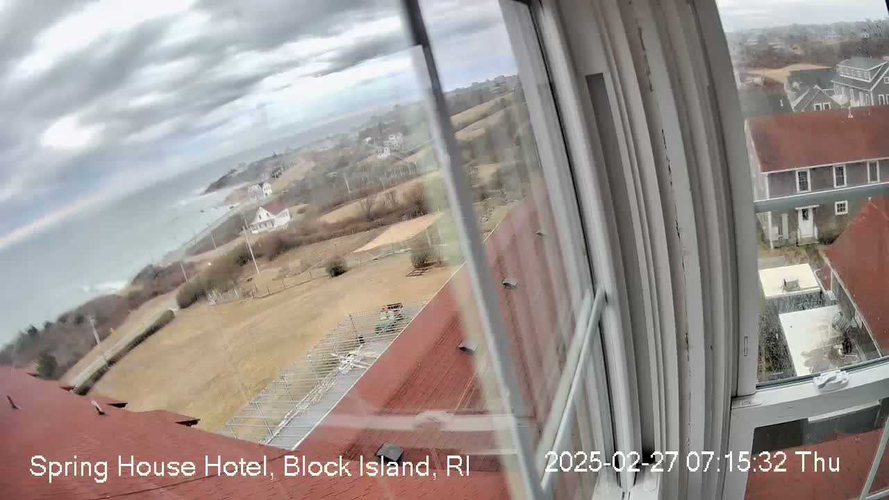 A view from a window shows a cloudy day overlooking a coastal town with buildings, ocean, and a grassy area.