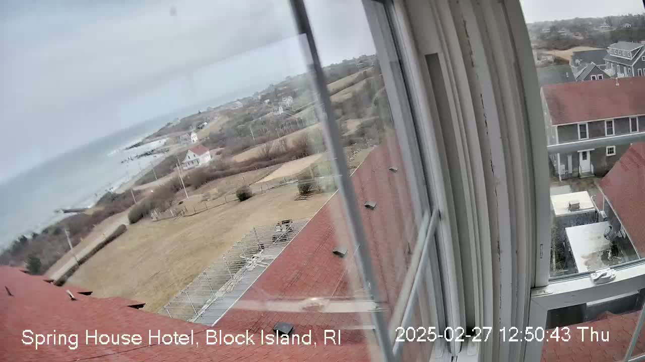 A view from a window shows a coastal town on an overcast day, with buildings, a grassy area, and the ocean visible in the distance.
