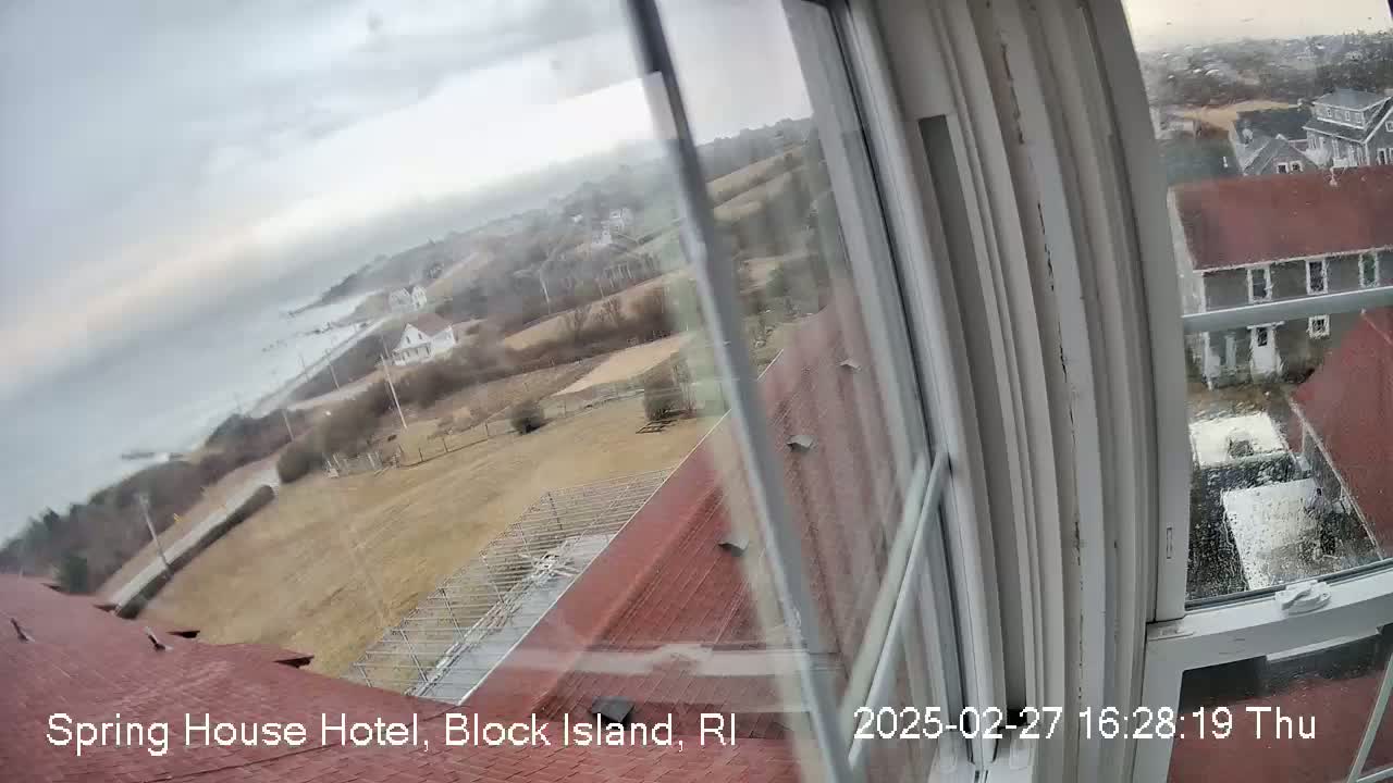 A view from a window shows a coastal scene under an overcast sky with a few buildings and a brown grassy area, partially obscured by rain on the windowpane.