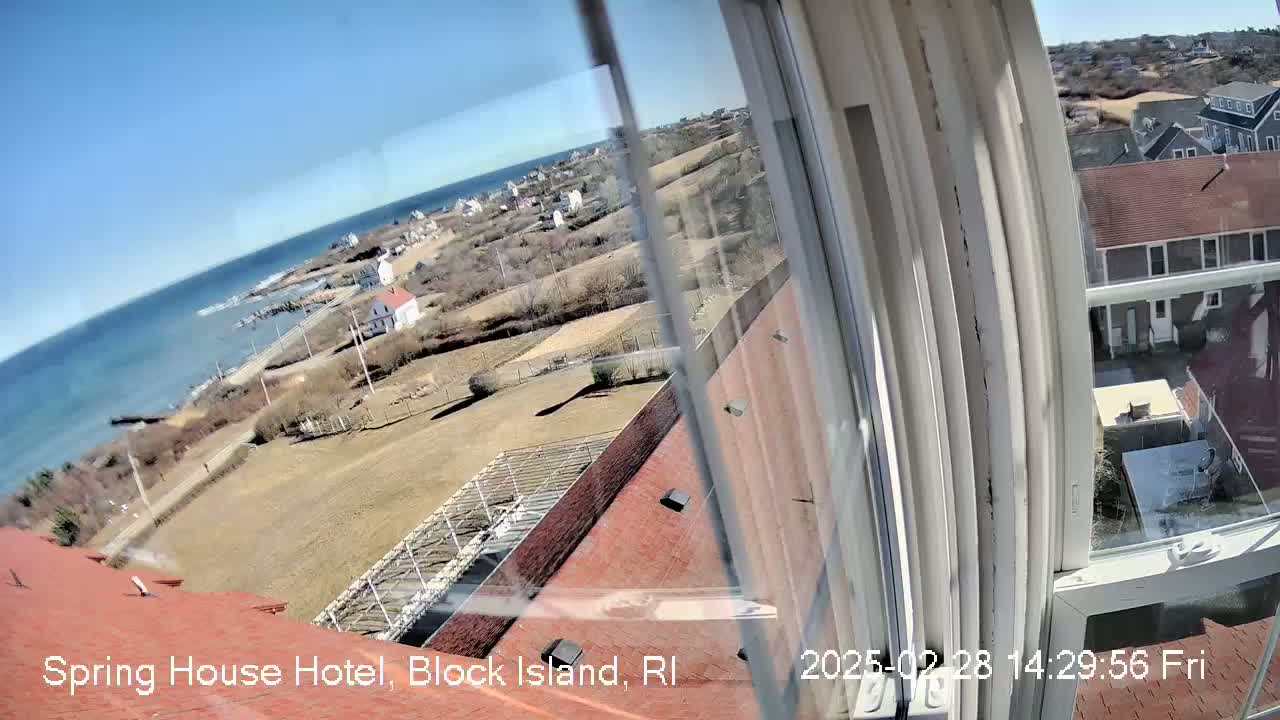 A view from a window shows a coastal town on a sunny day, with a body of water and rooftops visible.