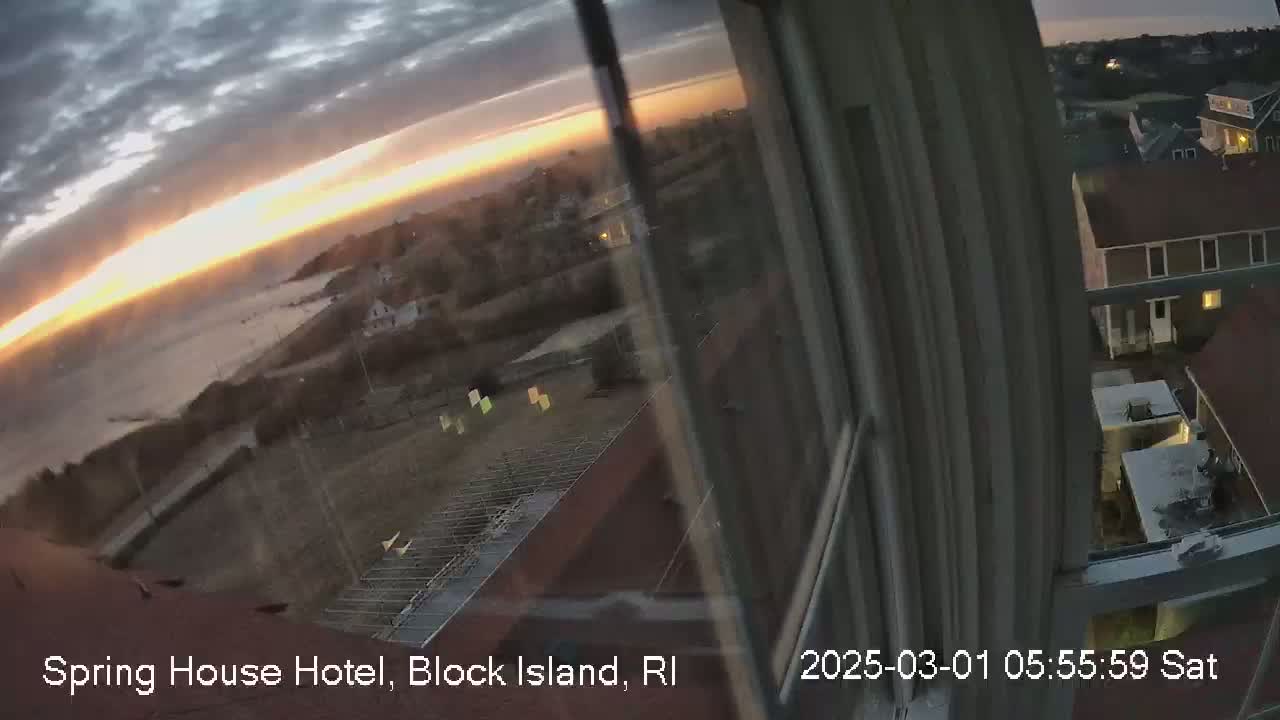 A sunrise over a calm ocean is viewed from a window overlooking a coastal town with some construction visible.