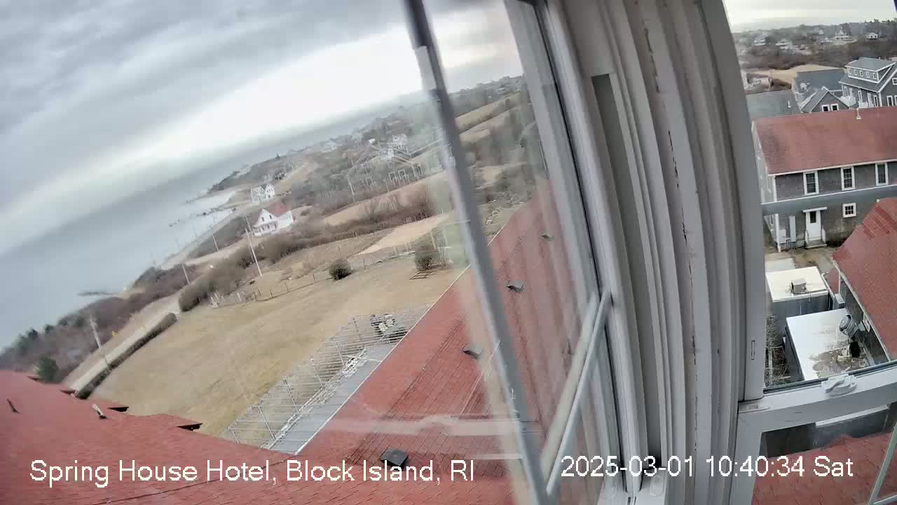 A view from a window shows a coastal landscape under an overcast sky, with buildings, a road, and the ocean visible in the distance.