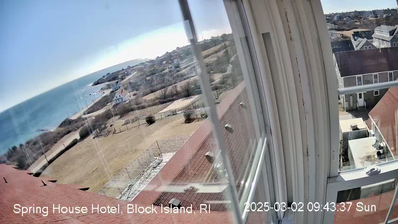 A sunny seaside view seen from a window shows a coastal town with buildings, a grassy area, and the ocean, partially obscured by the window frame.