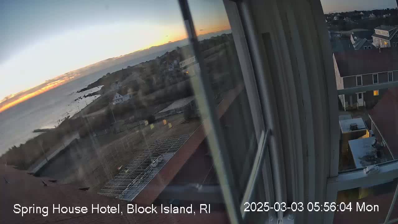 A sunrise over a calm ocean is viewed from a window, showing a coastal town with buildings and a partially constructed roof.