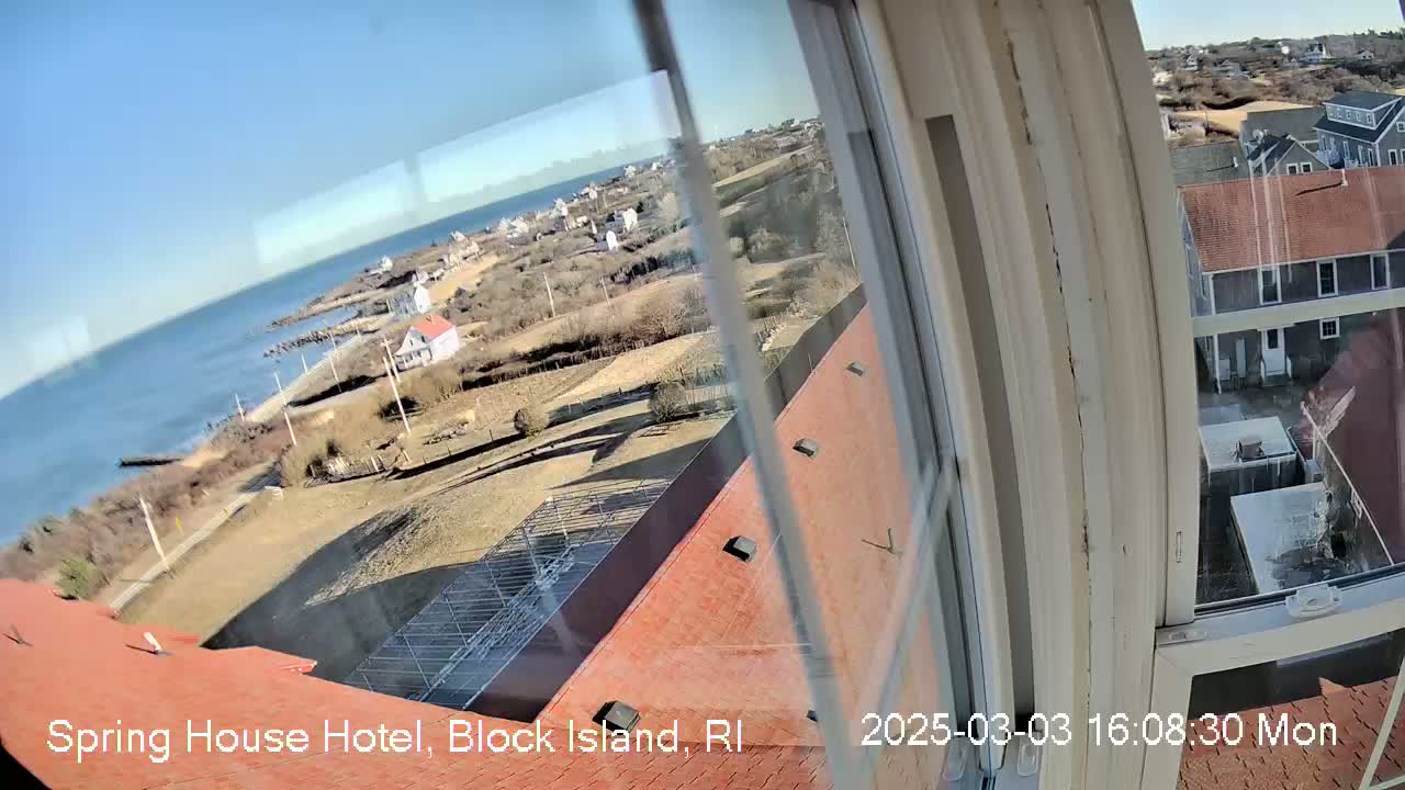 A view from a window shows a clear, sunny day overlooking a coastal town with red-roofed buildings, a body of water, and a sparsely vegetated landscape.