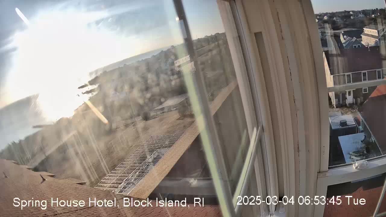 A view through a window shows a bright, sunny day overlooking a coastal town with a partially constructed structure in the foreground.