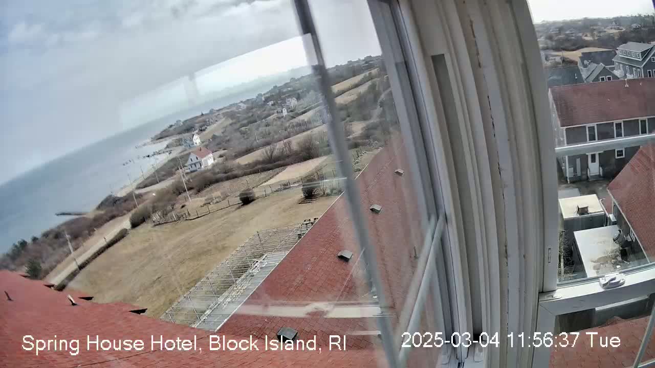 A view from a window shows a cloudy day overlooking a coastal town with brown rooftops and a body of water.