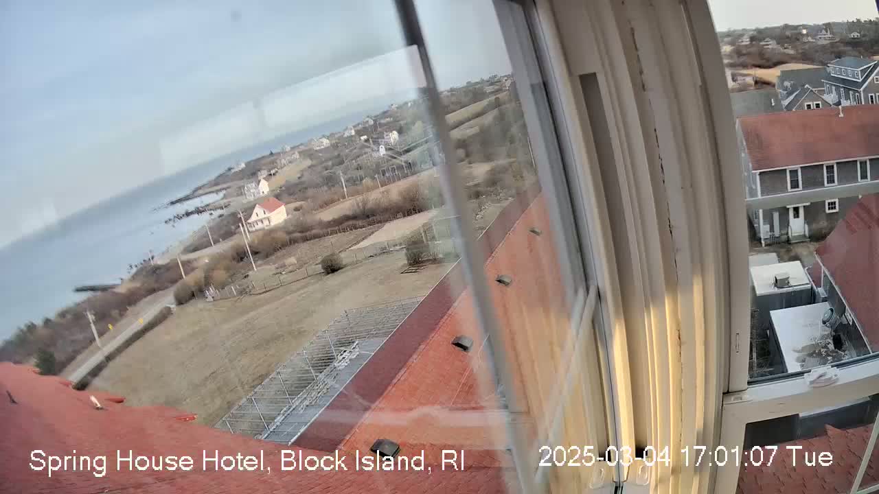 A high-angle view from a window shows a coastal town on a partly cloudy day, with a body of water visible in the distance.