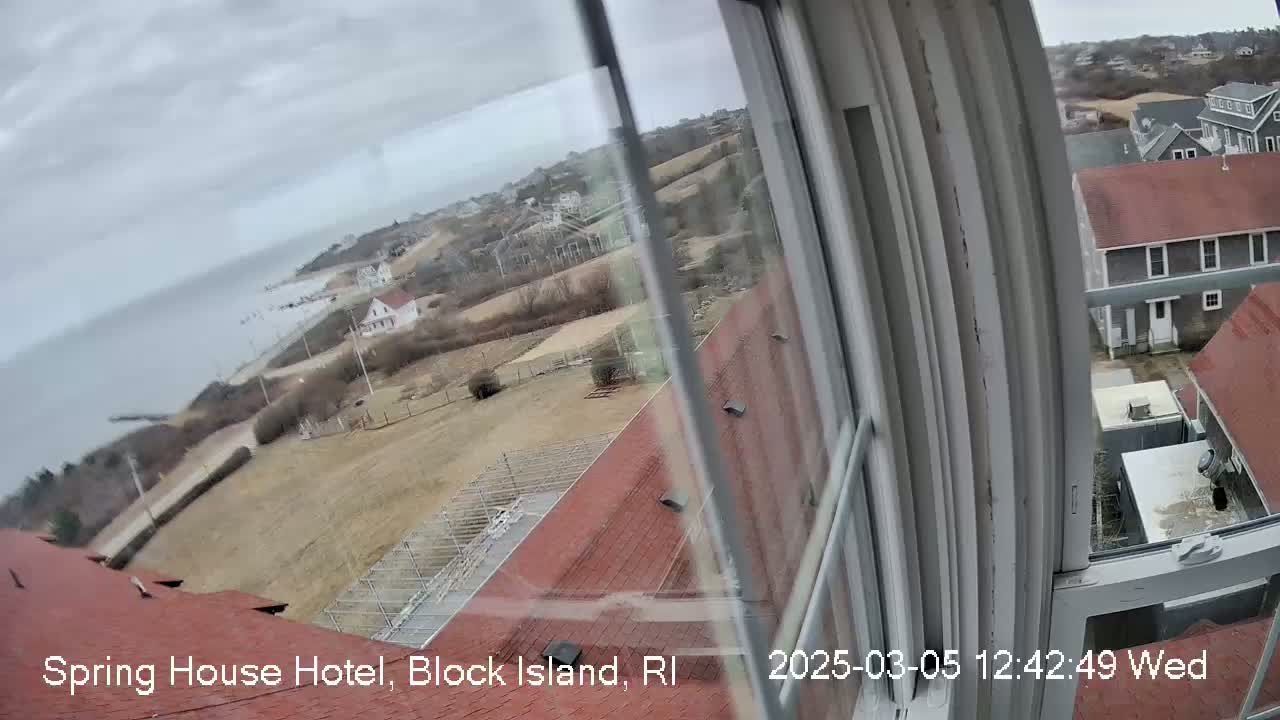 A view from a window shows a coastal town on an overcast day, with a body of water visible in the distance and buildings with red roofs in the foreground.