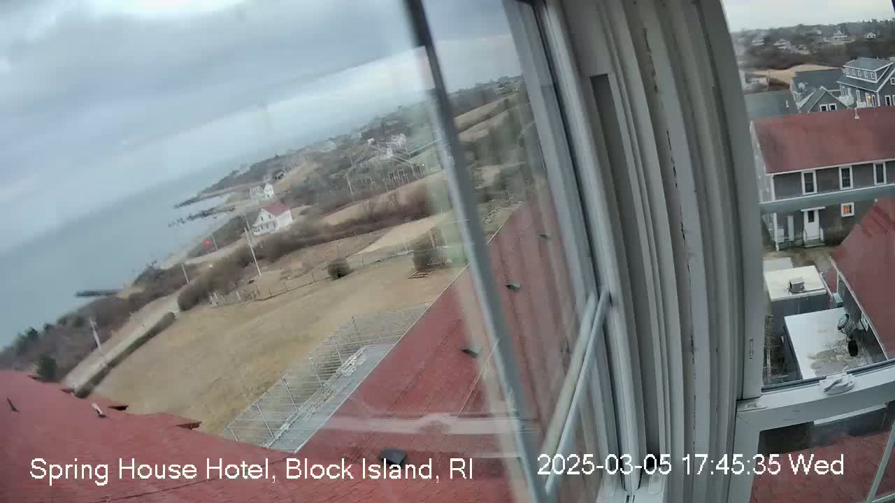 A view from a window shows a cloudy coastal landscape with buildings and a body of water under an overcast sky.