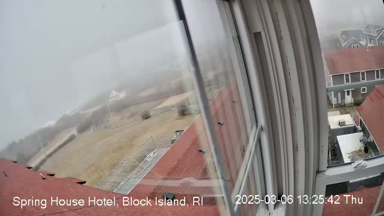 A foggy view from a window shows a red-roofed building, a fenced area, and other structures in the distance.