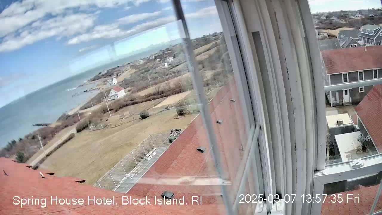 A partly cloudy day view from a window shows a coastal scene with buildings, grassy areas, and a body of water, seen from a high vantage point.