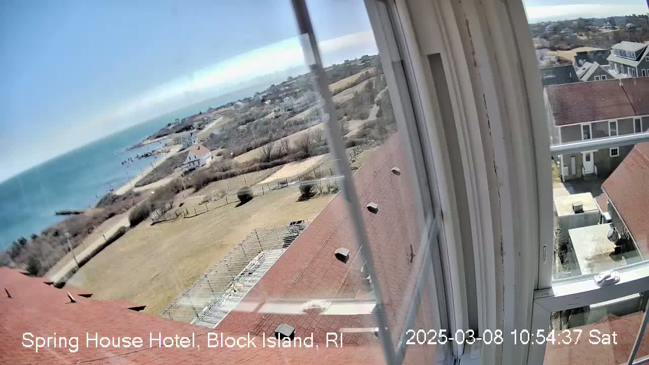 A view from a window shows a sunny coastal town with buildings, a shoreline, and ocean under a clear blue sky.