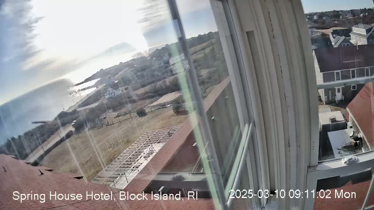 A view from a window shows a partly sunny day overlooking a coastal town with a body of water, buildings, and open land.