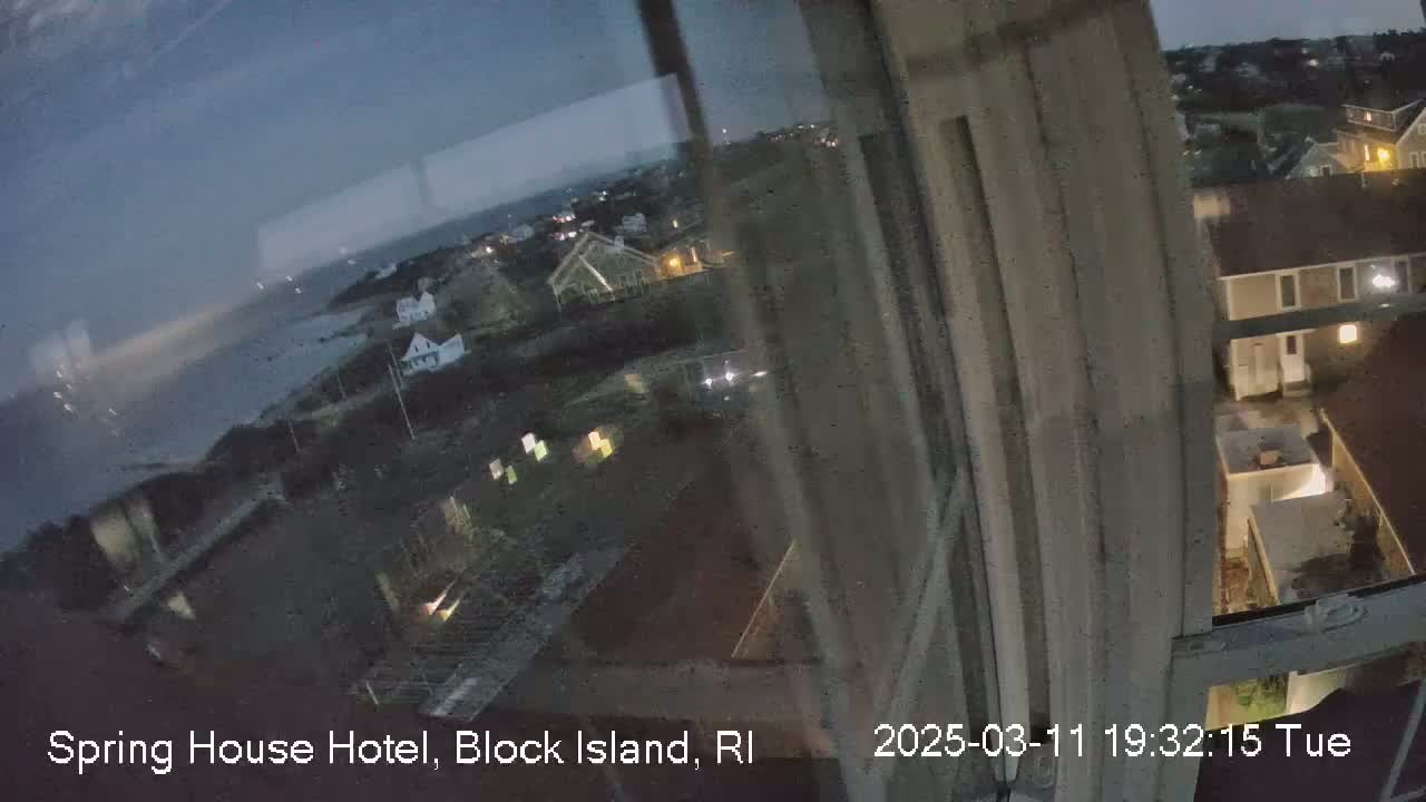 A nighttime view from a window shows a dark ocean, a coastline with houses, and a few lights visible in the distance under a dusky sky.