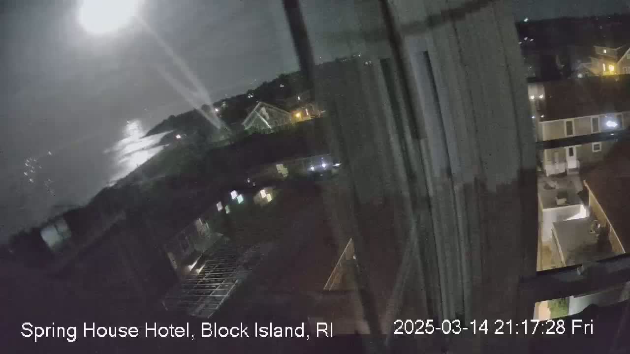 A nighttime, blurry view from a window shows a moonlit ocean and a residential area with houses, some with illuminated windows, under a cloudy sky.