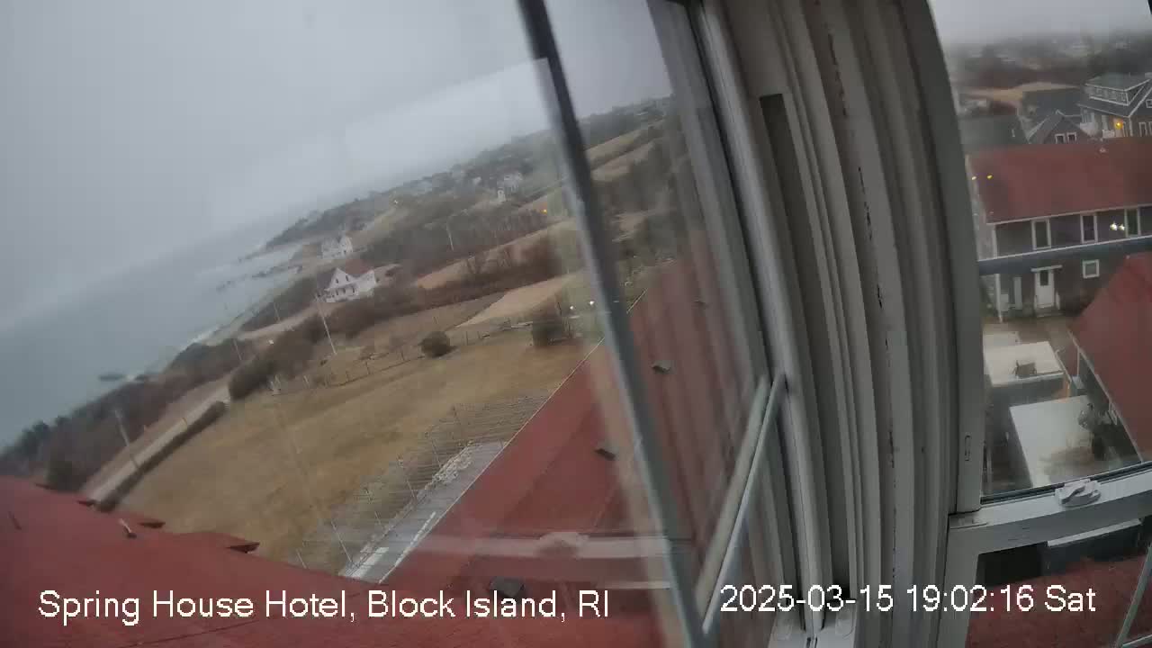 A view from a window shows an overcast coastal scene with houses and brown, grassy fields, extending to a grey ocean.