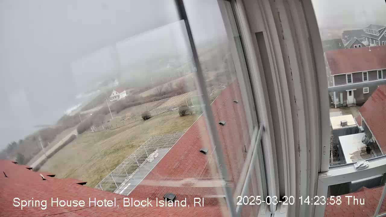 A foggy view from a window shows a red-roofed building, a grassy area with some structures, and a coastline in the distance.