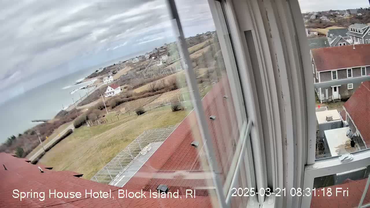 A view from a window shows a cloudy day overlooking a coastal town with red-roofed houses, a greenhouse, and a body of water.