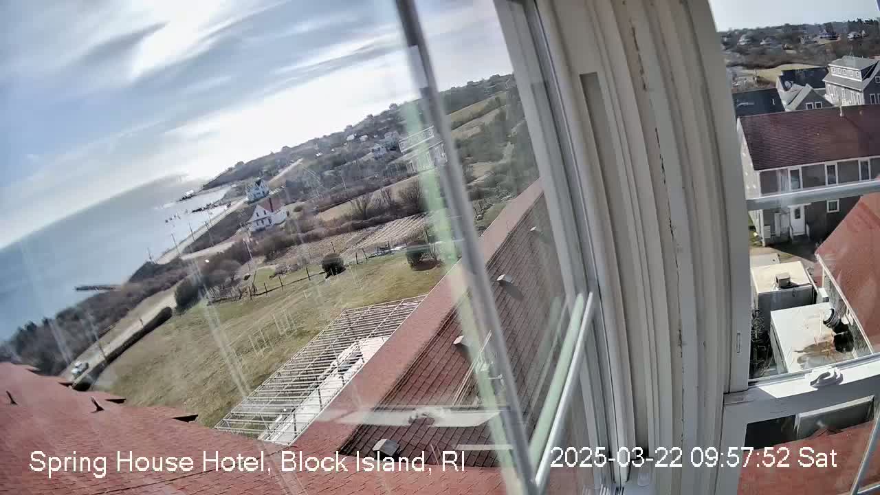 A view from a window shows a partly sunny day overlooking a coastal town with buildings, a road, and ocean, seen through a slightly dirty window.