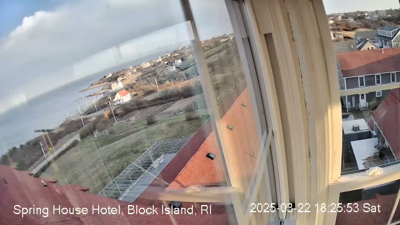A view from a window shows a partly cloudy day over a coastal town with buildings, open fields, and a body of water.