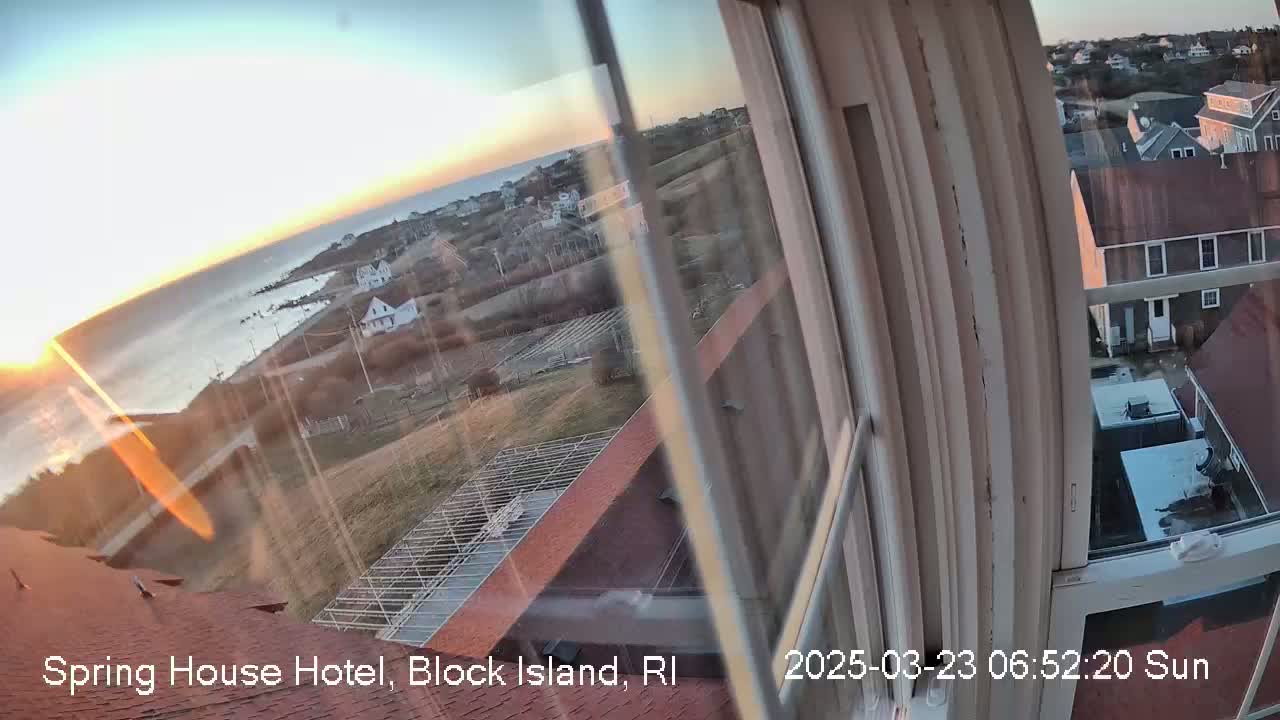 A sunrise over a calm ocean is viewed from a window, showing a coastal town with houses and a red roof below.