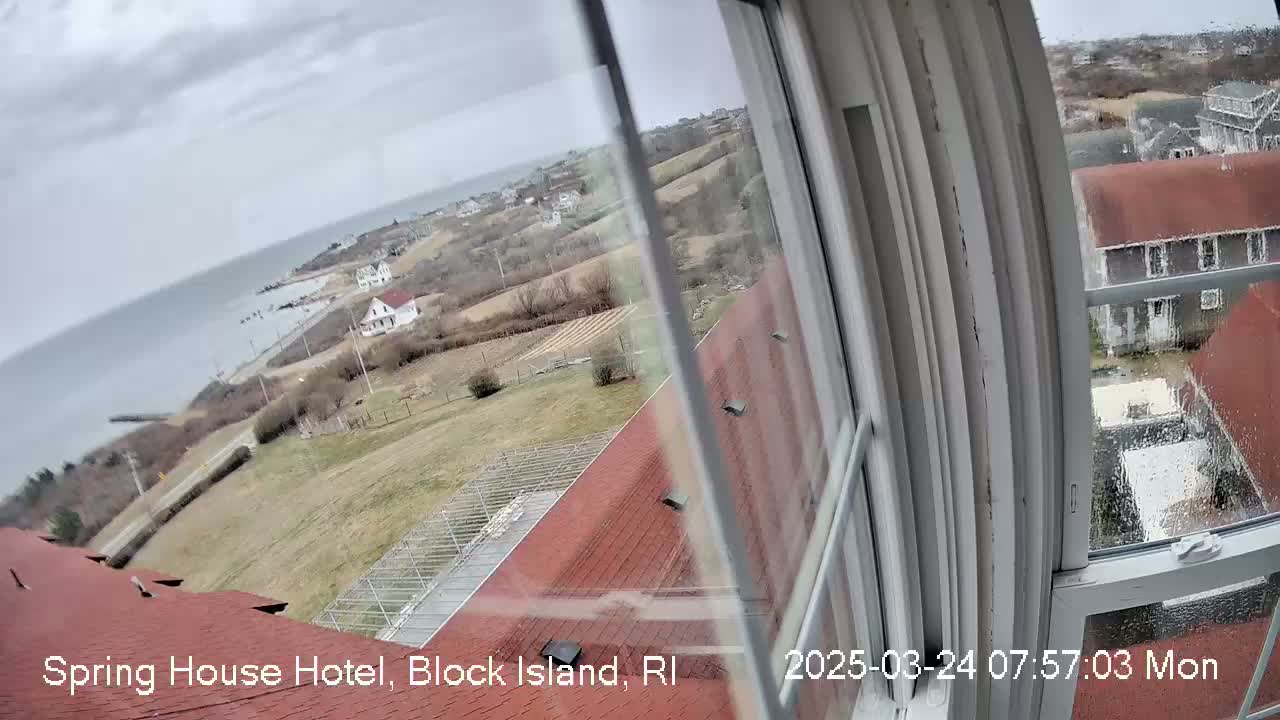 A view from a window shows a coastal landscape under an overcast sky, with houses, fields, and the ocean visible in the distance.