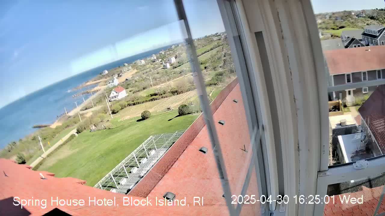 A sunny view from a window shows a red roof, a grassy lawn, several buildings, and a body of water extending to the horizon.
