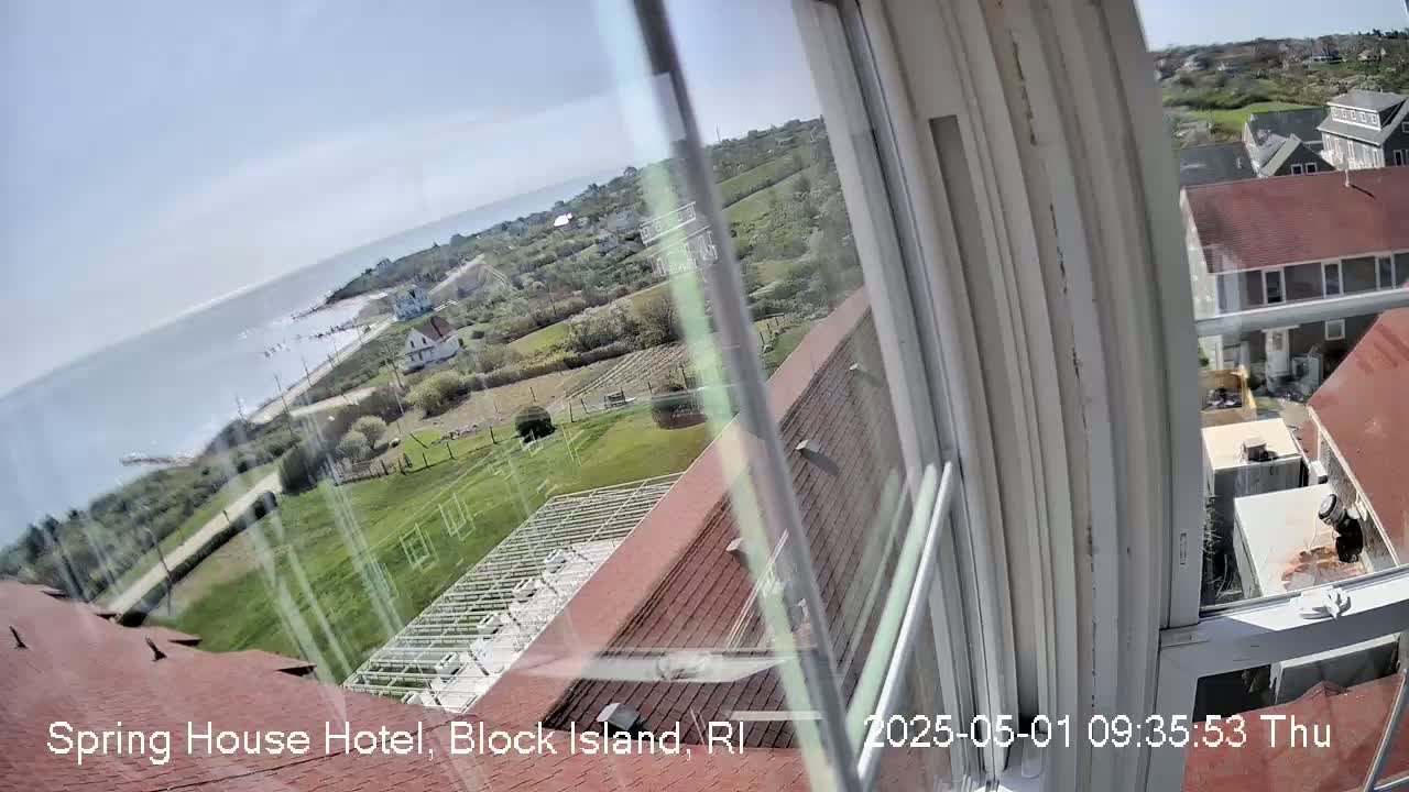 A view from a window shows a clear, sunny day over a coastal area with houses, green lawns, and the ocean in the distance.