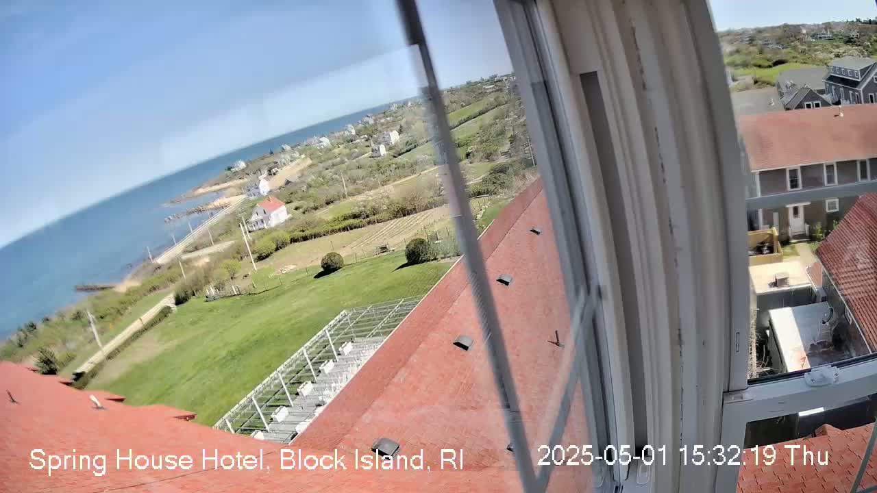 A sunny view from a window shows a red roof, green lawns, ocean, and coastal buildings.