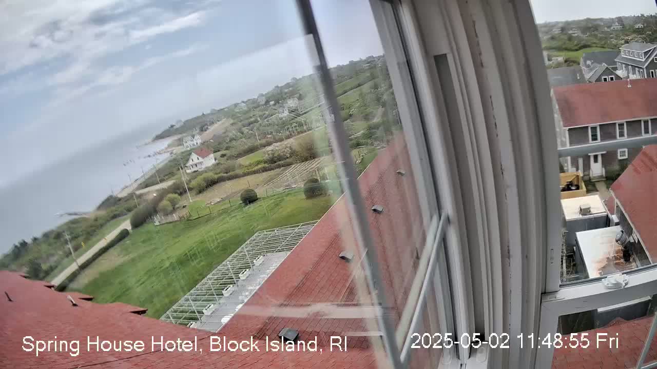 A view from a window shows a coastal town with red-roofed buildings under an overcast sky, overlooking a body of water.