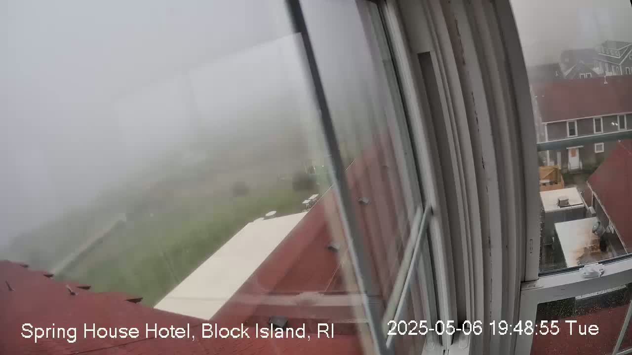 A foggy view from a window shows red rooftops and a grassy area, with additional buildings visible in the distance.