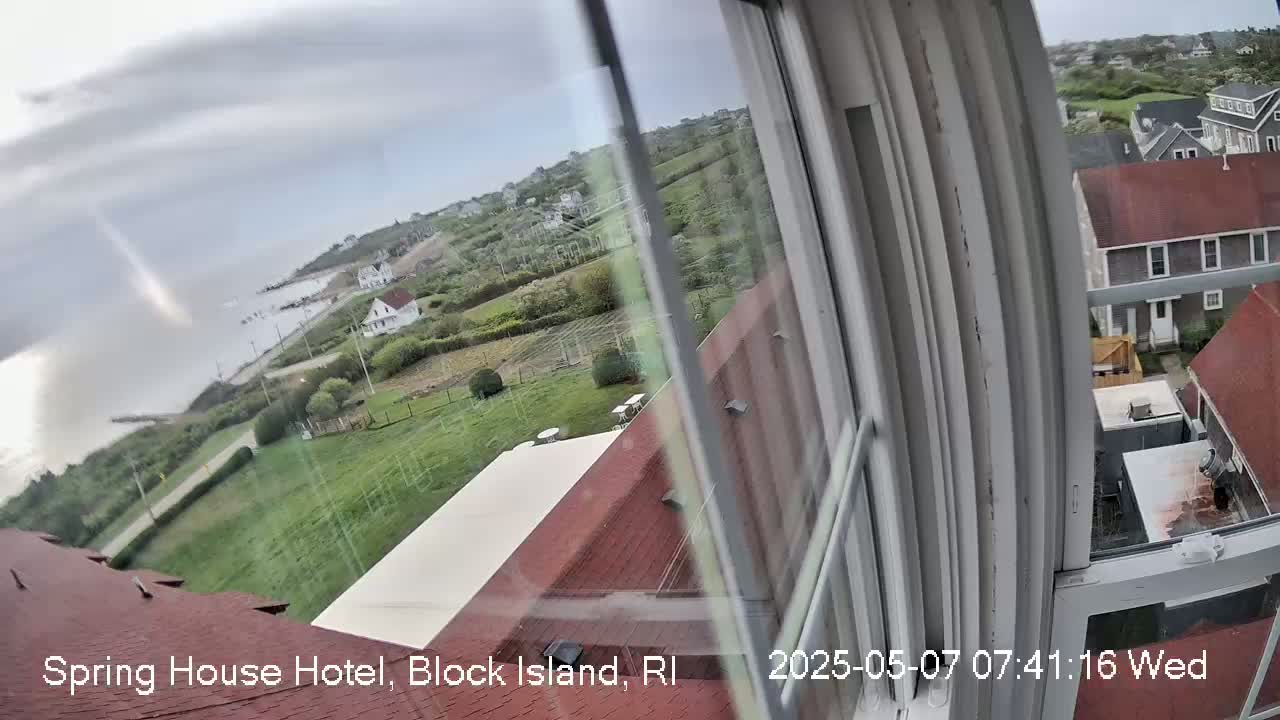 A view from a window shows a cloudy day overlooking a coastal town with houses, green lawns, and a body of water.