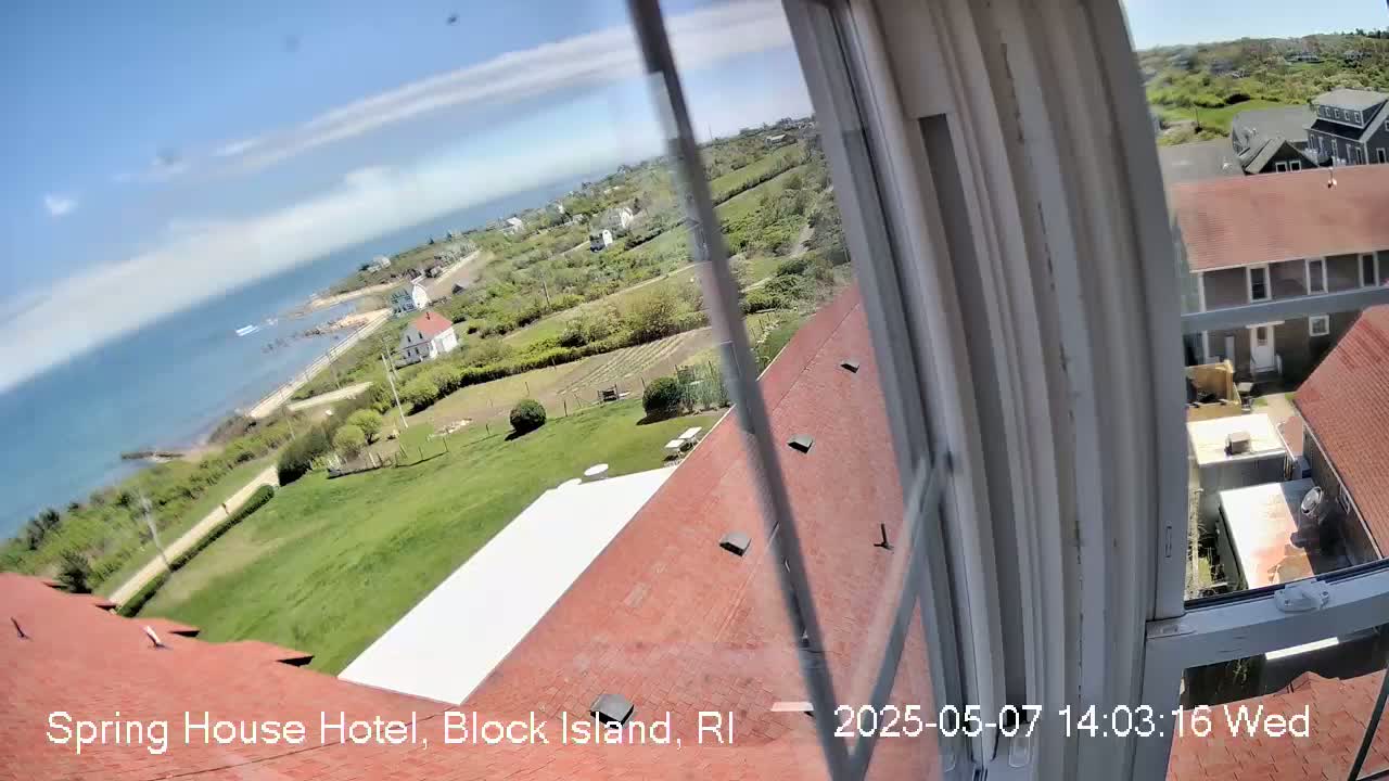 A view from a window shows a red roofed building overlooking a coastal town with green lawns, houses, and a body of water under a partly cloudy sky.