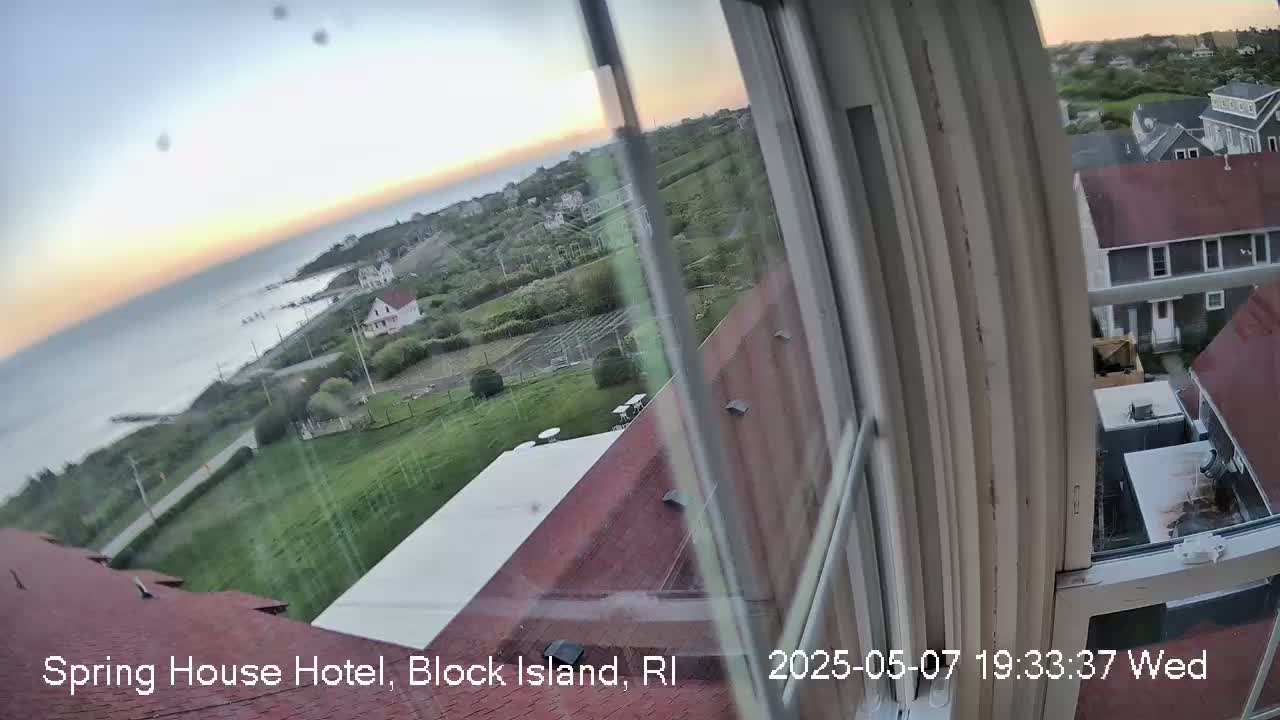 A view from a window shows a calm ocean at sunset, coastal buildings, and green lawns.