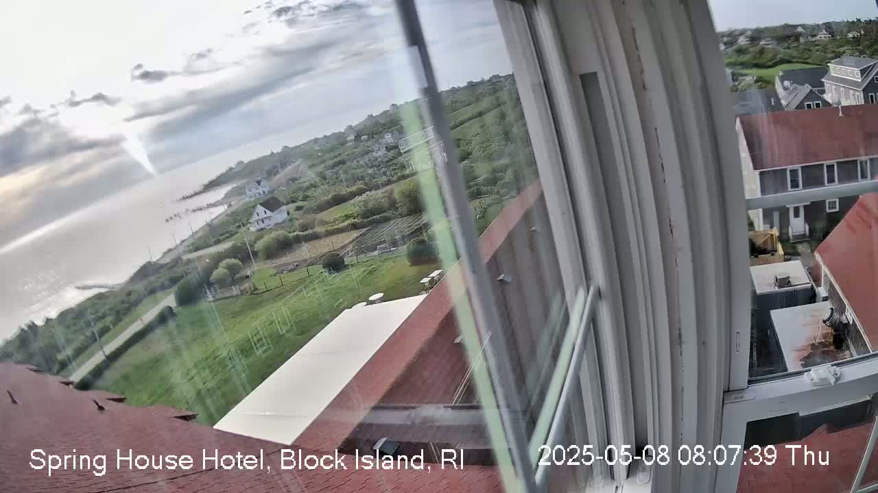 A partly cloudy view from a window shows a calm ocean, a coastal road, green lawns, and houses, with a red roof visible in the foreground.