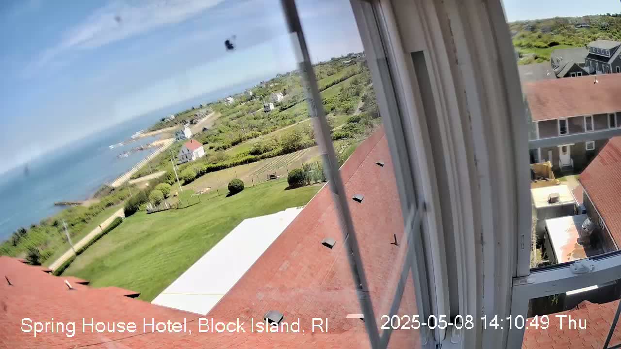 A view from a window shows a red roof, a grassy lawn, several buildings, and a blue ocean under a sunny sky.