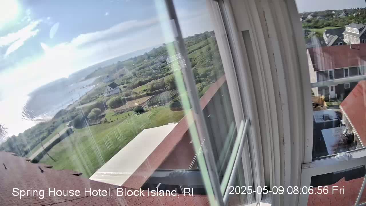A view from a window shows a partly sunny day overlooking a coastal area with houses, green lawns, and the ocean in the distance.