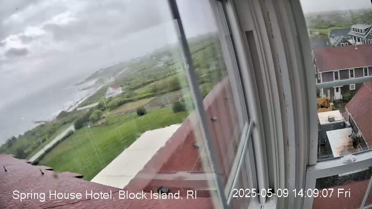 A view from a window shows a rain-swept coastal landscape with green fields, buildings, and a gray ocean under an overcast sky.