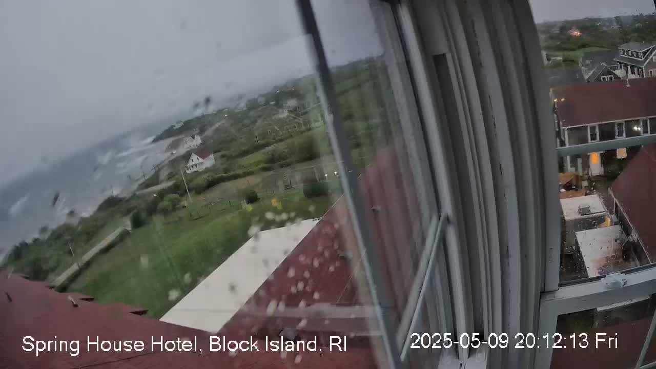 A rainy view from a window shows a coastal scene with houses, green spaces, and a stormy ocean.