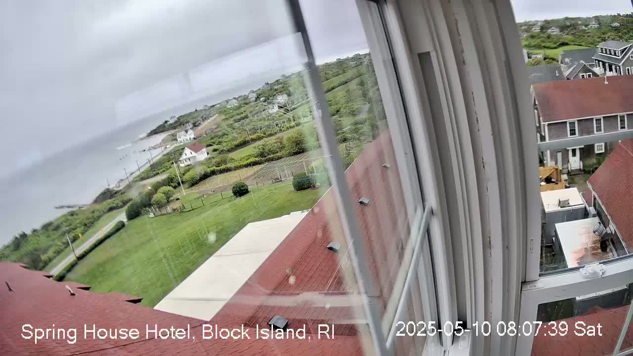 A view from a window shows an overcast sky over a coastal area with green lawns, houses, and the ocean in the distance.