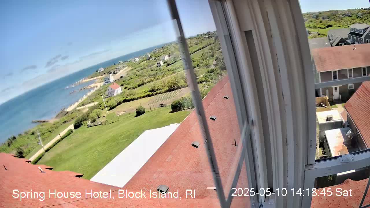 A view from a window shows a coastal landscape with a red-roofed building, green lawns, and a body of water under a clear blue sky.