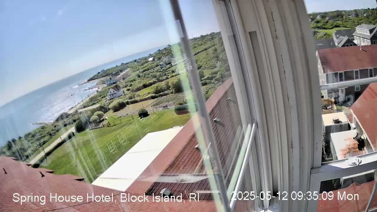 A view from a window shows a partly sunny day overlooking a coastline with a body of water, green lawns, and buildings.