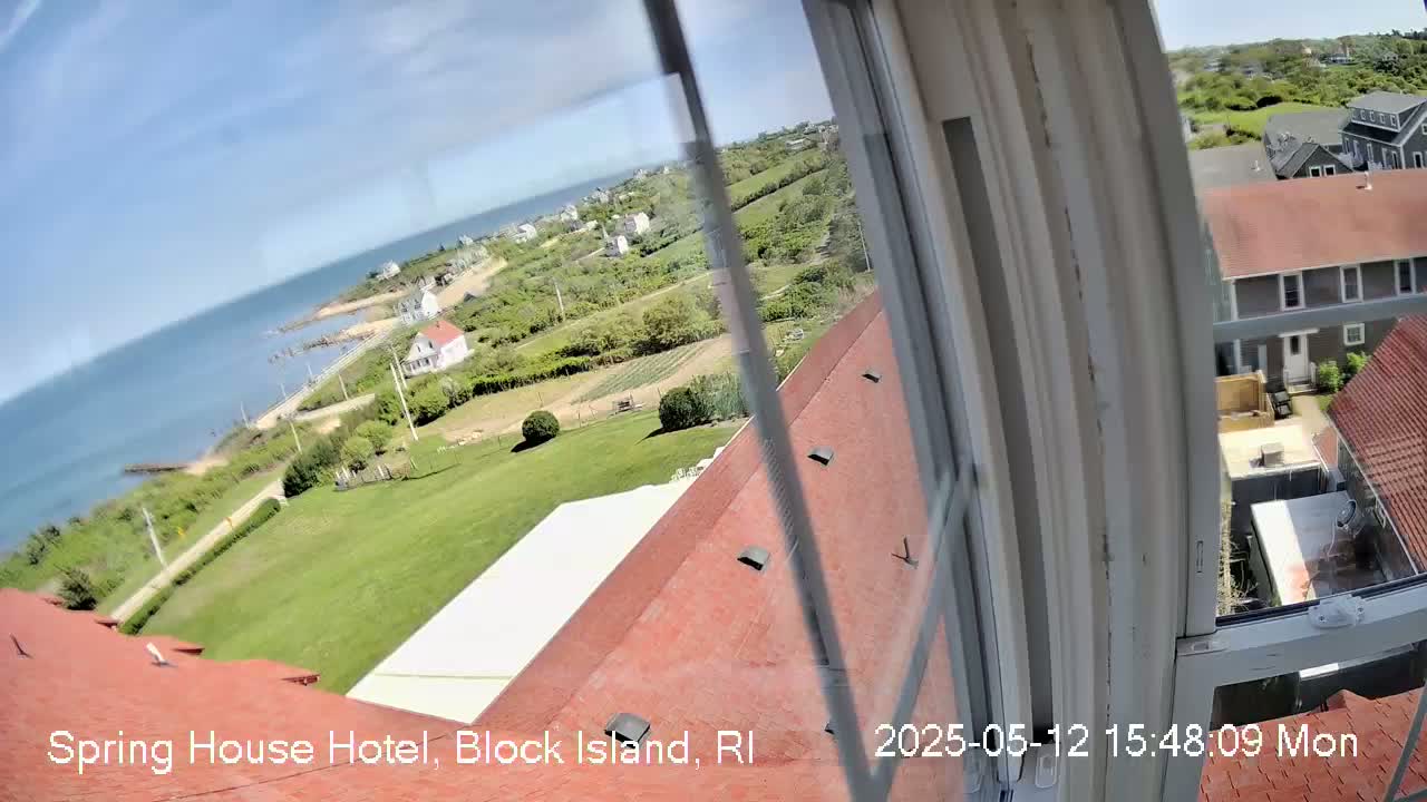 A view from a window shows a sunny coastal scene with a red roofed building in the foreground, green lawns and houses, and a blue ocean in the distance.